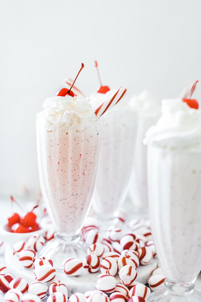 Three glasses filled with Chick Fil A peppermint milkshake. There are tiny bowls of cherries to the side and peppermint candies scattered around the glasses.