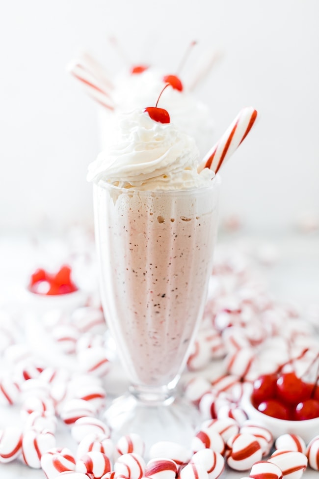 Chick Fil A peppermint milk shake in a tall glass topped with whipped cream, a cherry, and a peppermint stick. There are peppermint candies scattered around it.