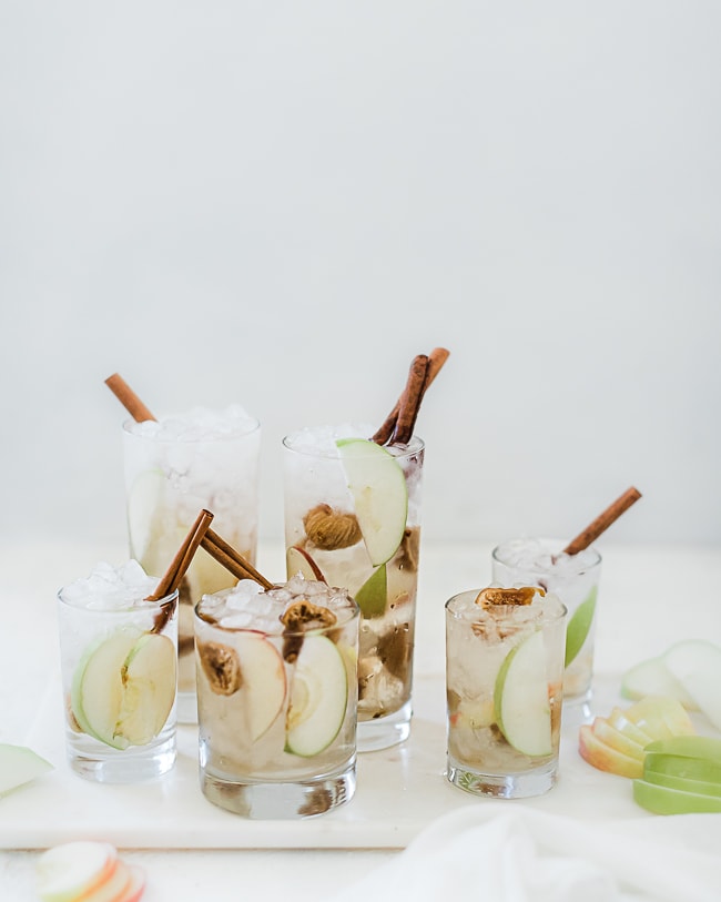 Glasses filled with apple pie fruit water recipe. Drinks are garnished with cinnamon sticks and there are apple slices scattered around.