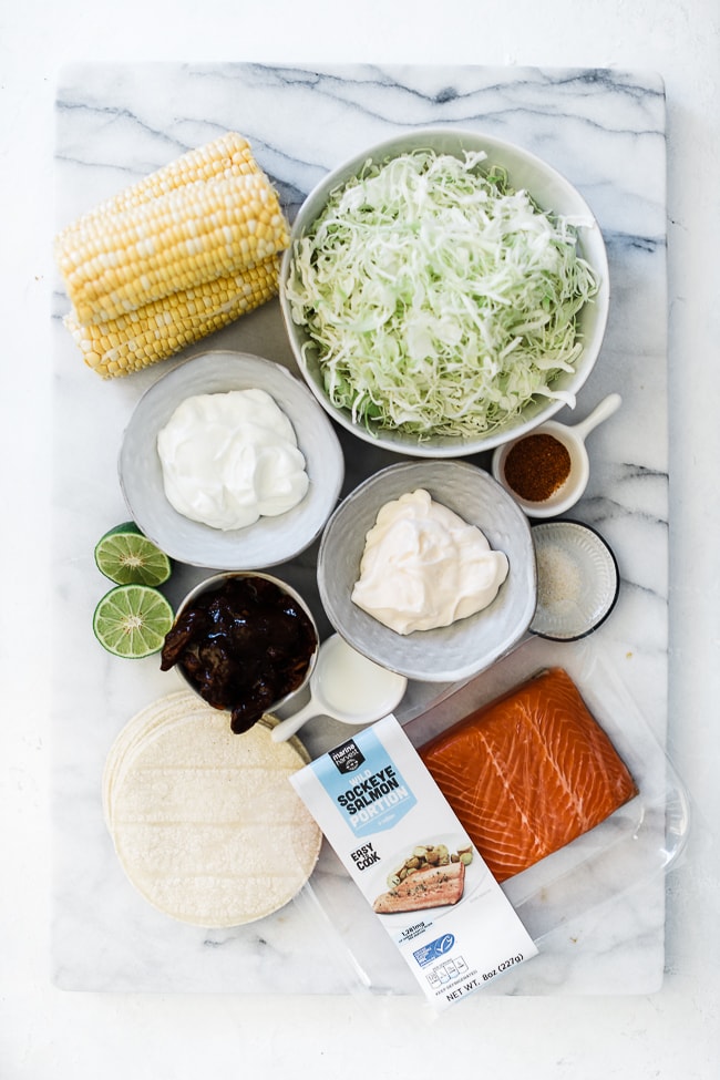 Corn, cabbage, sour cream. mayo, chipotle peppers, limes, salmon, and corn tortillas on a marble board.