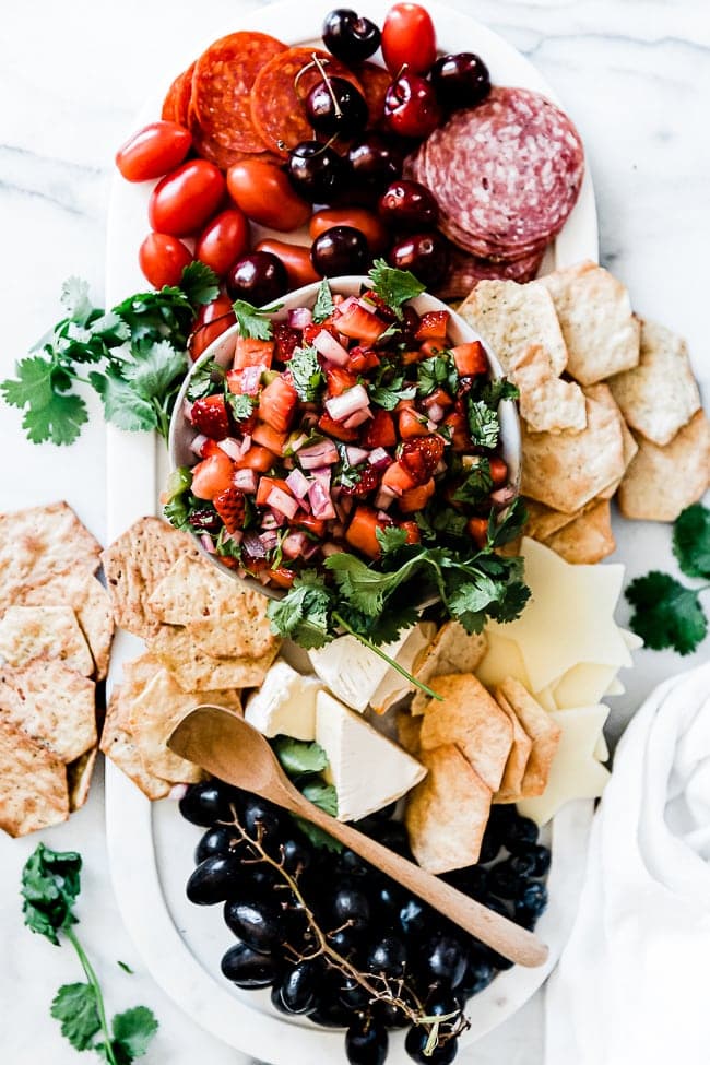 Strawberry salsa in a black rimmed bowl atop a marble platter. It is surrounded by crackers, meats, cheeses, and fruit.