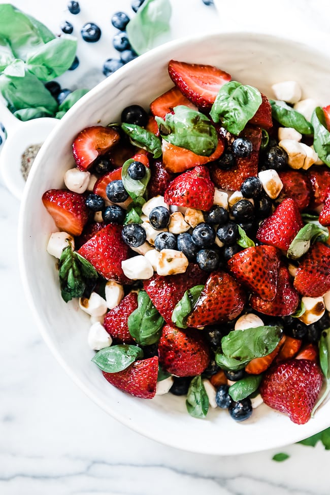 Summer strawberry salad in a white bowl. There is basil and blueberries to the side.