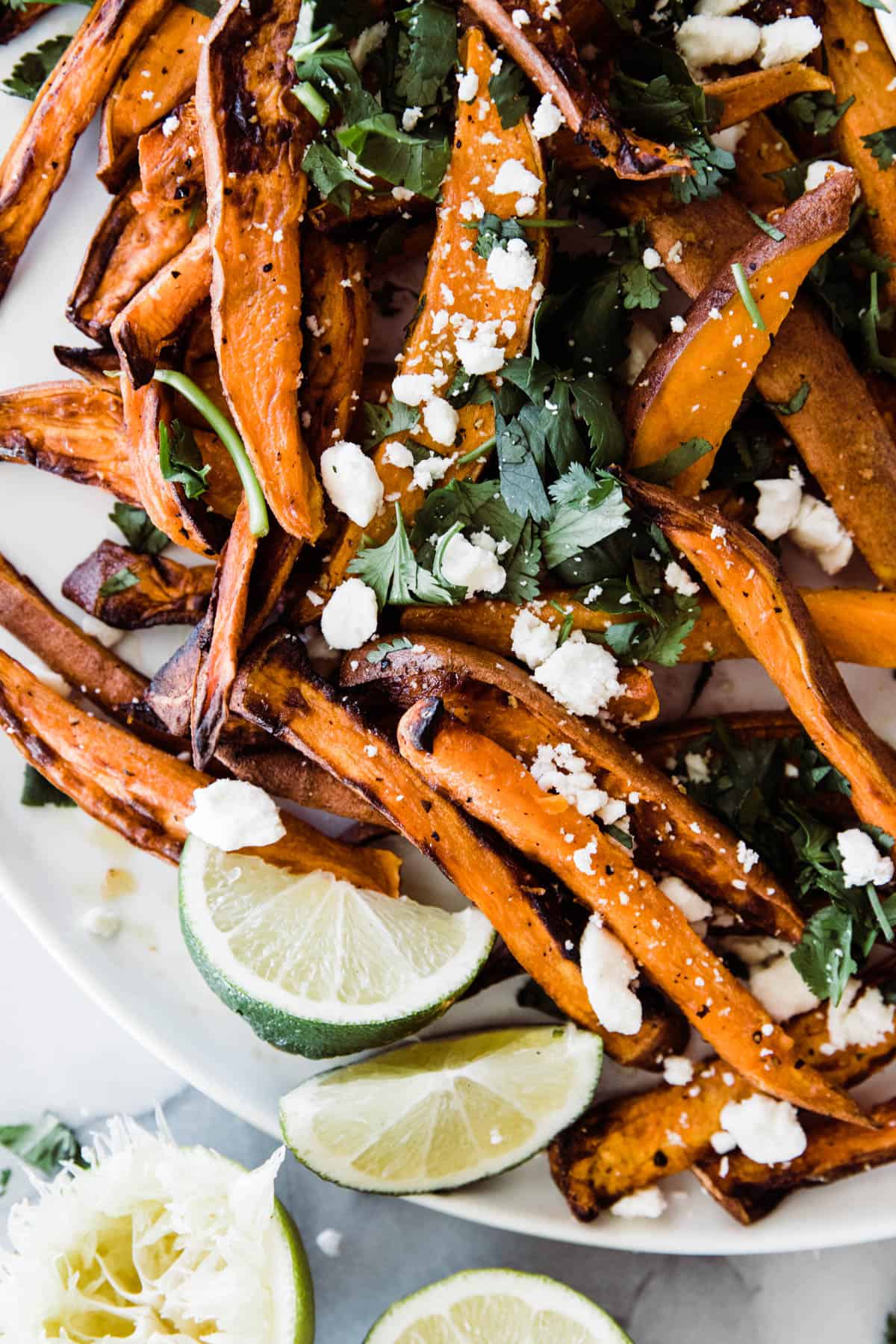 White plate of sweet potato fries with two dipping sauces, and some limes as garnishes and feta cheese on top. 