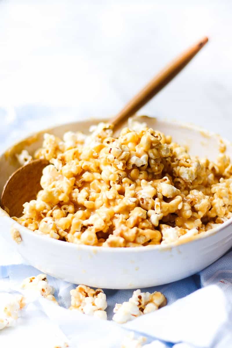 A side shot of caramel popcorn in a white bowl with a wooden serving spoon