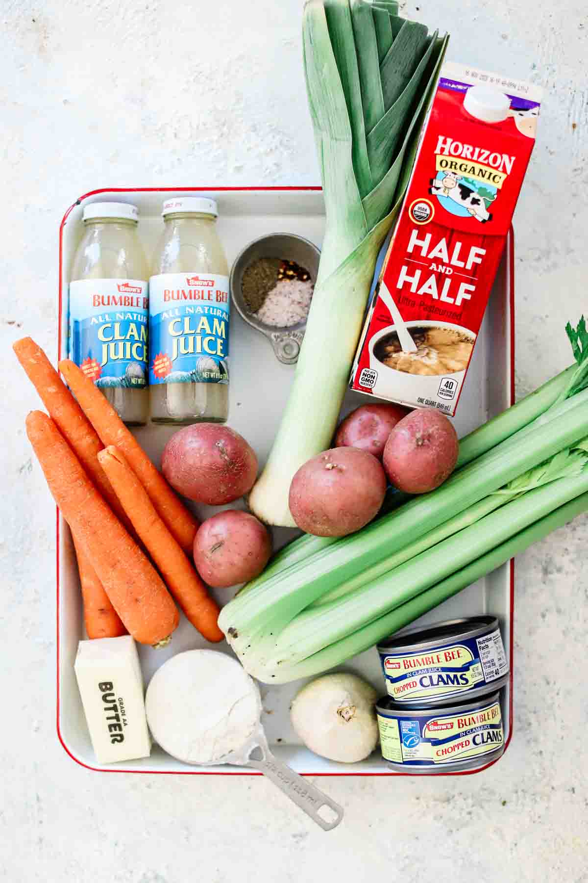 The ingredients needed for clam chowder on a baking sheet including clam juice, potatoes, carrots, celery, flour, butter, clams, cream, and leeks.