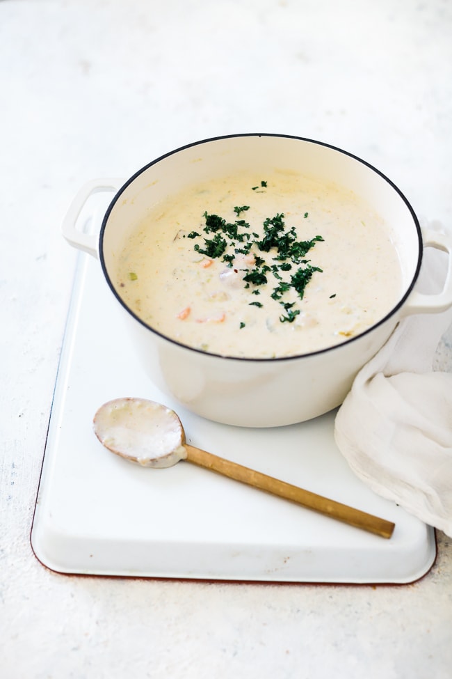 Boston clam chowder in a white dutch over. The cast iron pot is atop a white baking sheet and a wooden spoon is set to the side.
