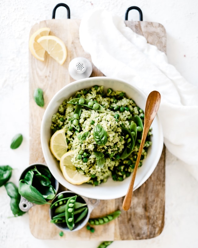 Lemon pesto cauliflower rice recipe in a white bowl, garnished with peas and lemon slices. Bowl is set on top of a wooden cutting board.