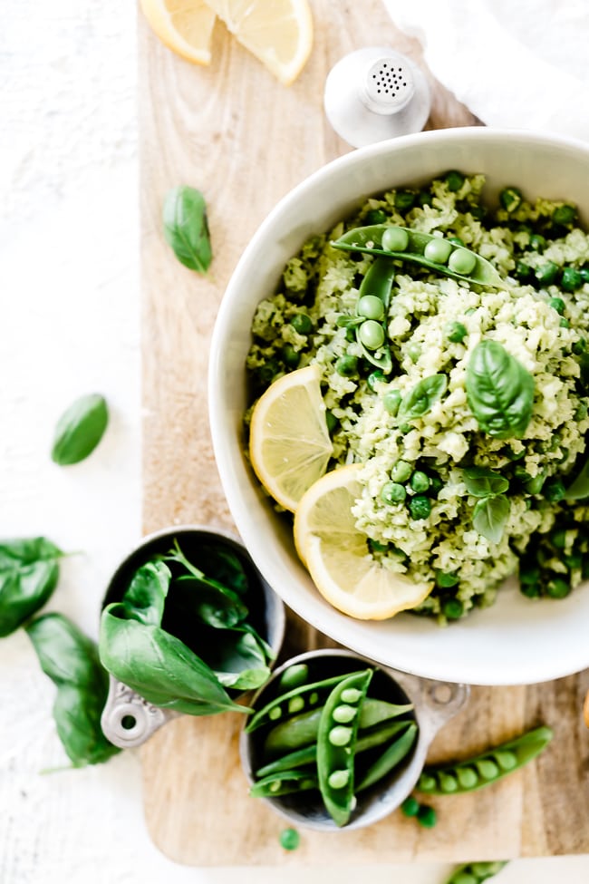 Lemon pesto cauliflower rice recipe in a white bowl, garnished with peas and lemon slices. Bowl is set on top of a wooden cutting board.