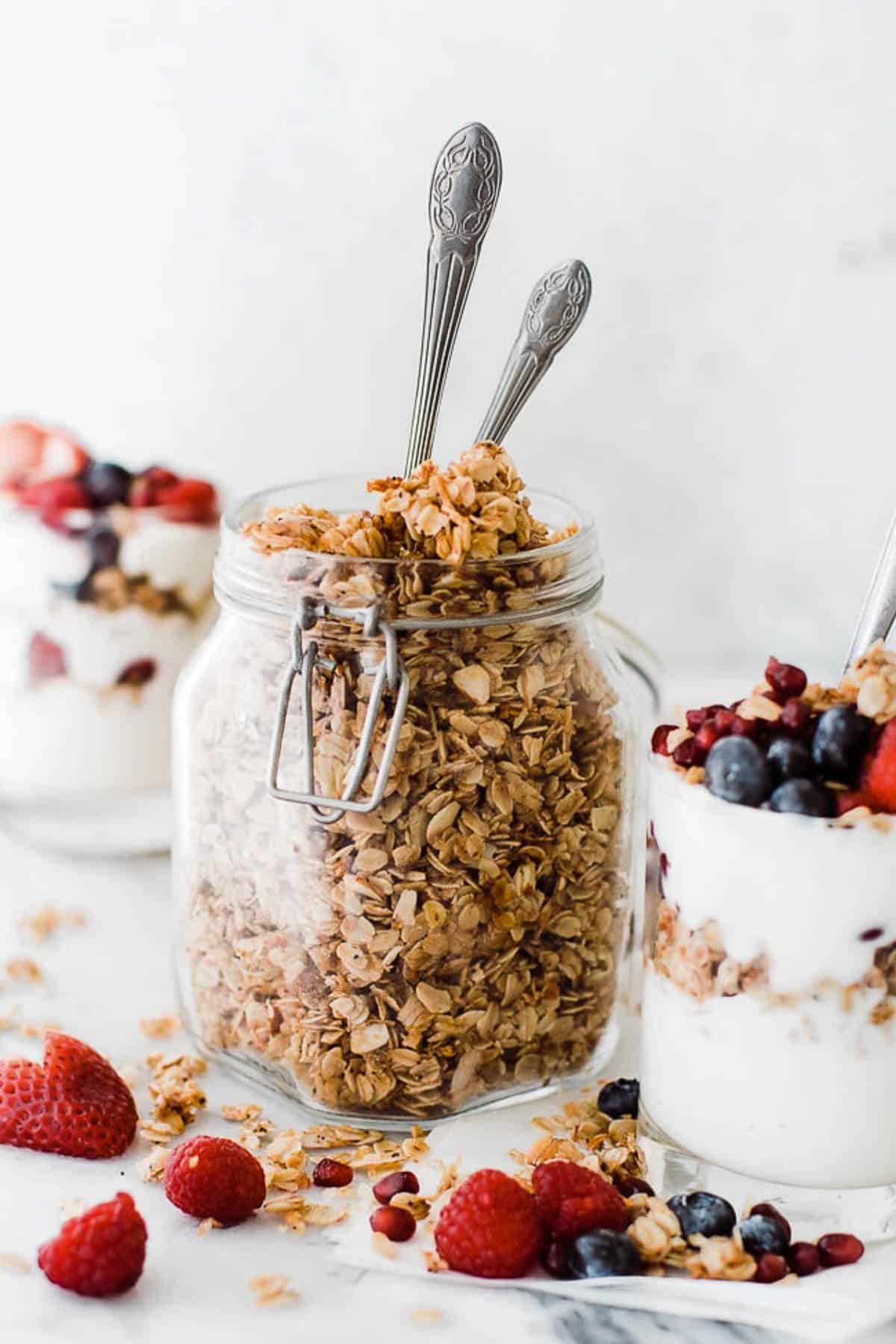 A jar of healthy granola on the table with two spoons in it and yogurt and berries in a glass.