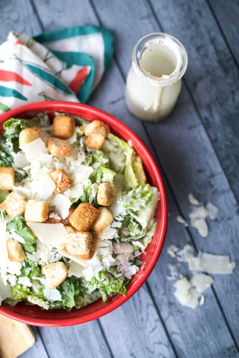 Caesar Dressing in a little jar next to a hearty salad in a big red bowl topped with homemade croutons 