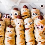 several mummy dogs (hot dogs wrapped in bread and have eyes) laying on a tray