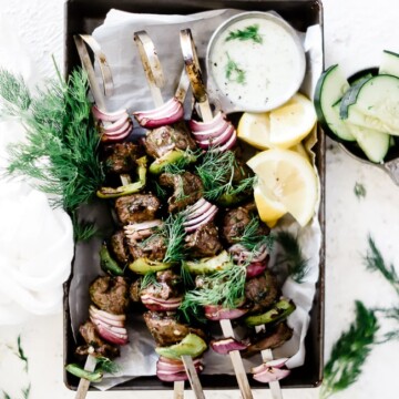 Lemon dill lamb kabobs in a baking tray, garnished with fresh dill and a side of tzatziki.