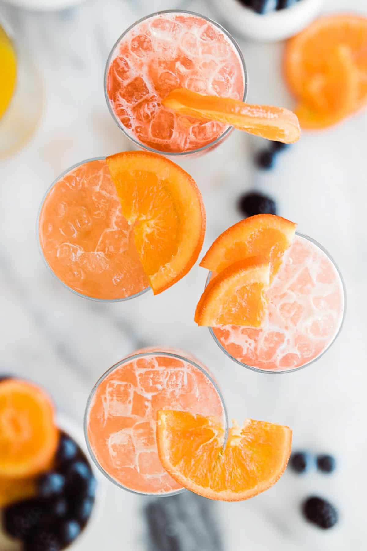 An overhead shot of four glasses of virgin mimosas with cranberry juice garnished with oranges on the table.