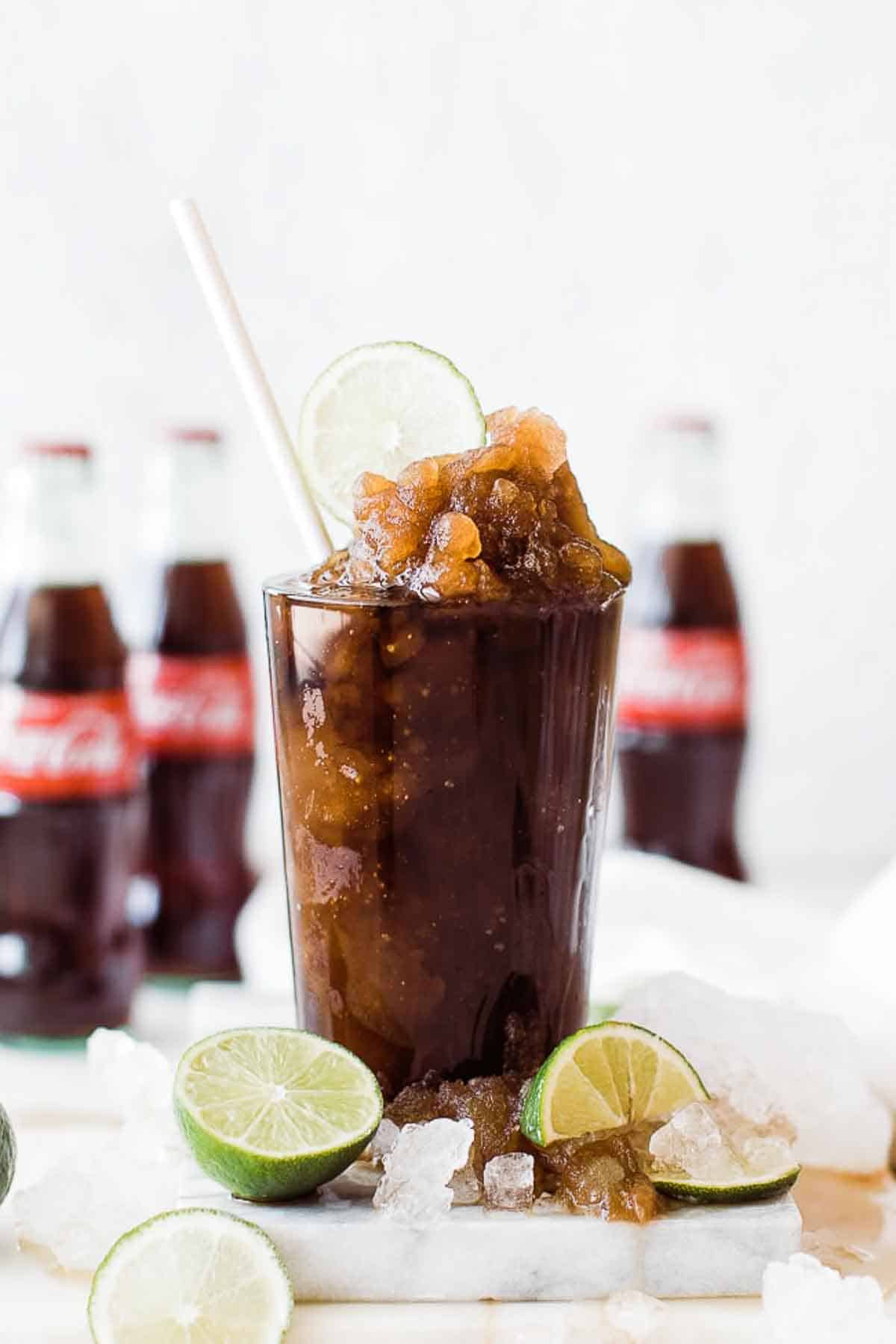 A glass of copycat Coke slurpee on the table with crushed ice and limes and bottles of Coke in the background.