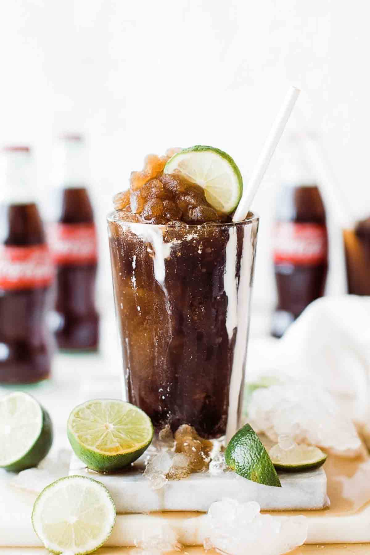 A frozen coke slushie in a glass garnished with a lime and a straw.