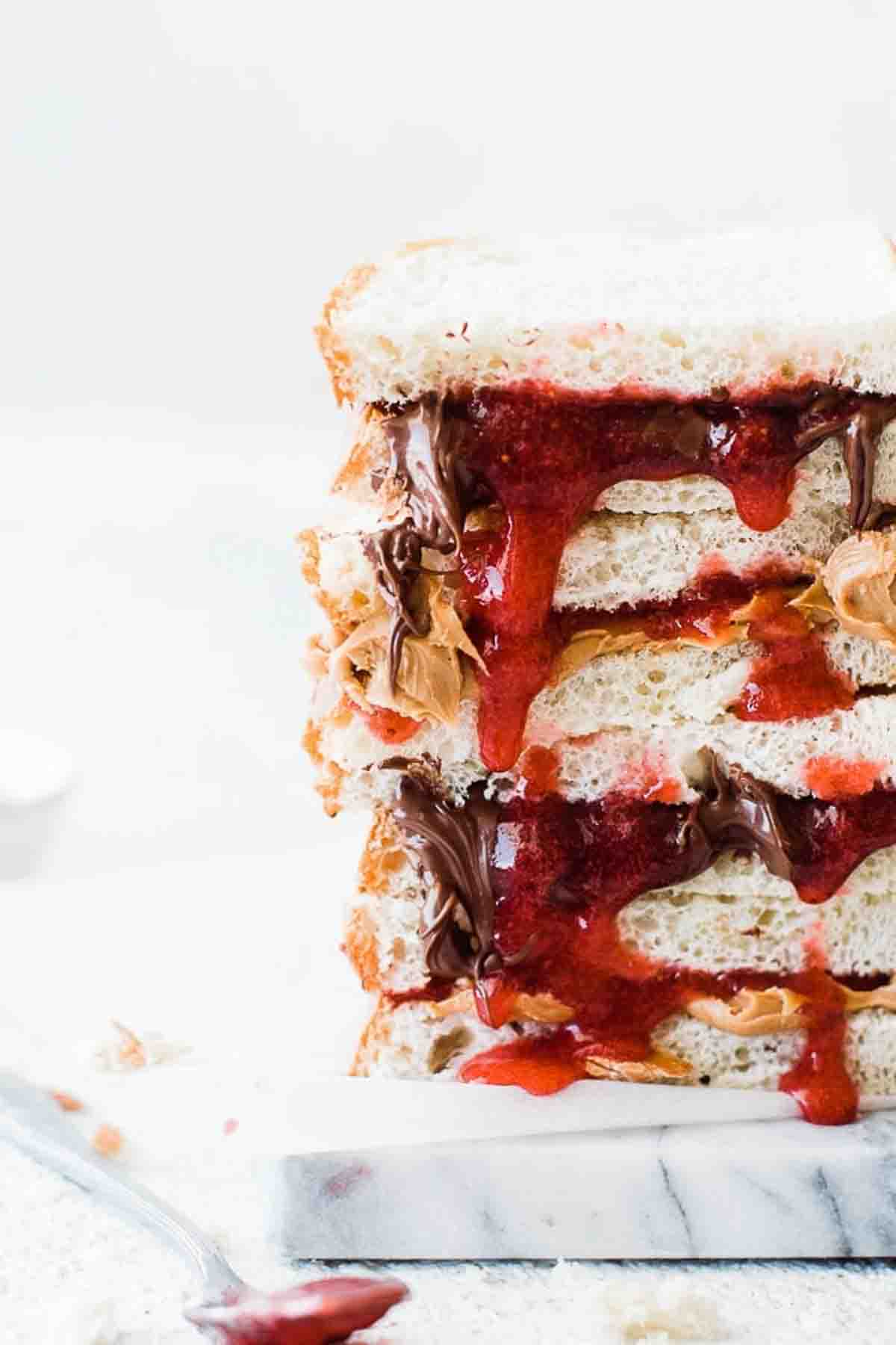 Close up of sandwiches stacked on top of each other with homemade freezer jam dripping down and nutella and peanut butter. inbetween