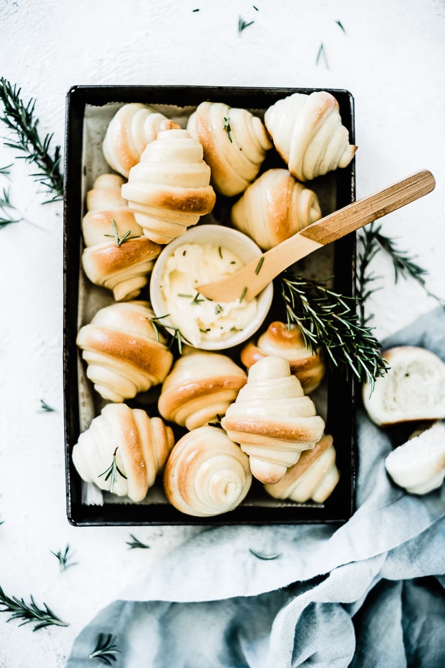 Homemade crescent rolls on a vintage baking pan with a small bowl of butter and rosemary sprigs.