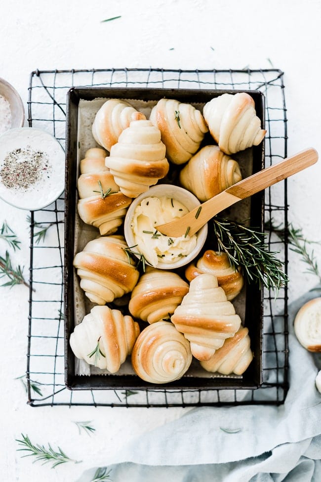 Homemade crescent rolls on a vintage baking pan with a small bowl of butter and rosemary sprigs.