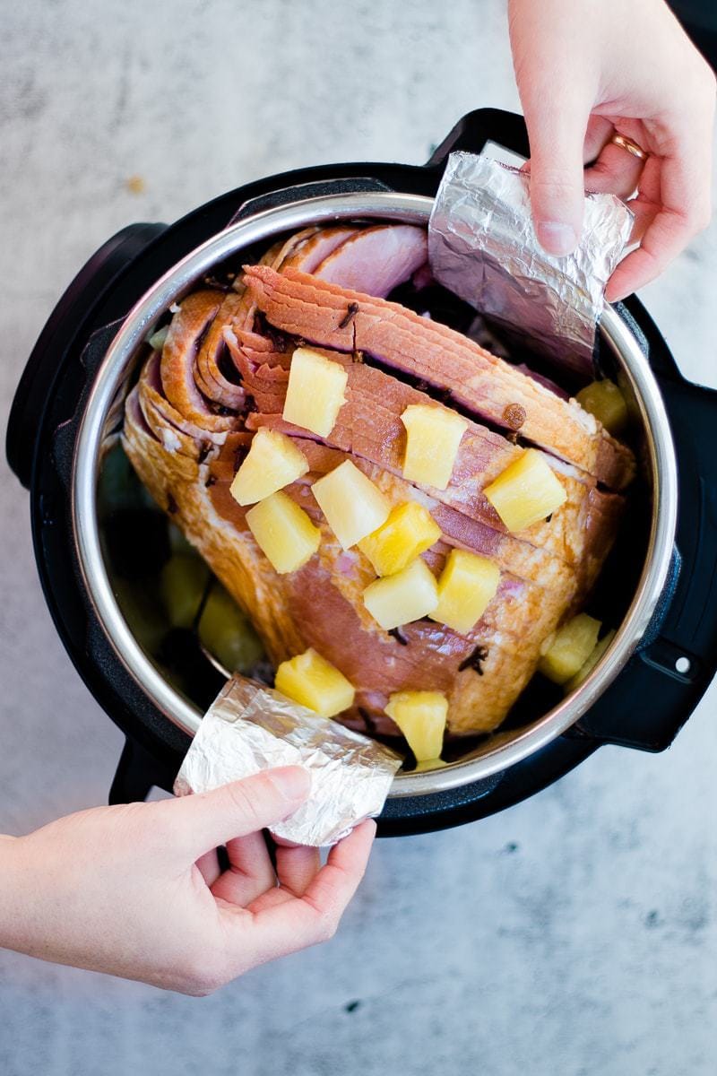 An overhead shot of pressure cooker ham in a pressure cooker with chunks of pineapple and demonstration of foil handles