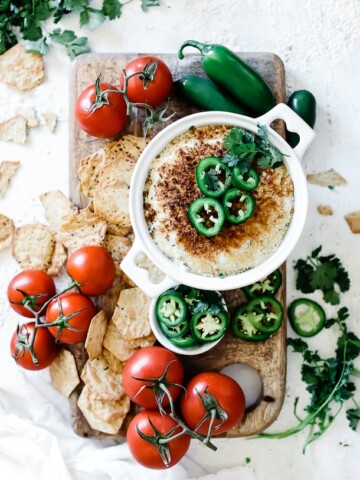 Jalapeno popper dip recipe in a round white baking dish. The dish is placed atop a wooden board and is surrounded by crackers and fresh tomatoes. The dip is garnished with jalapeños.