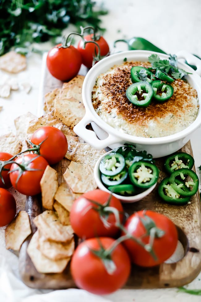 Jalapeno popper dip recipe in a round white baking dish. The dish is placed atop a wooden board and is surrounded by crackers and fresh tomatoes. The dip is garnished with jalapeños.