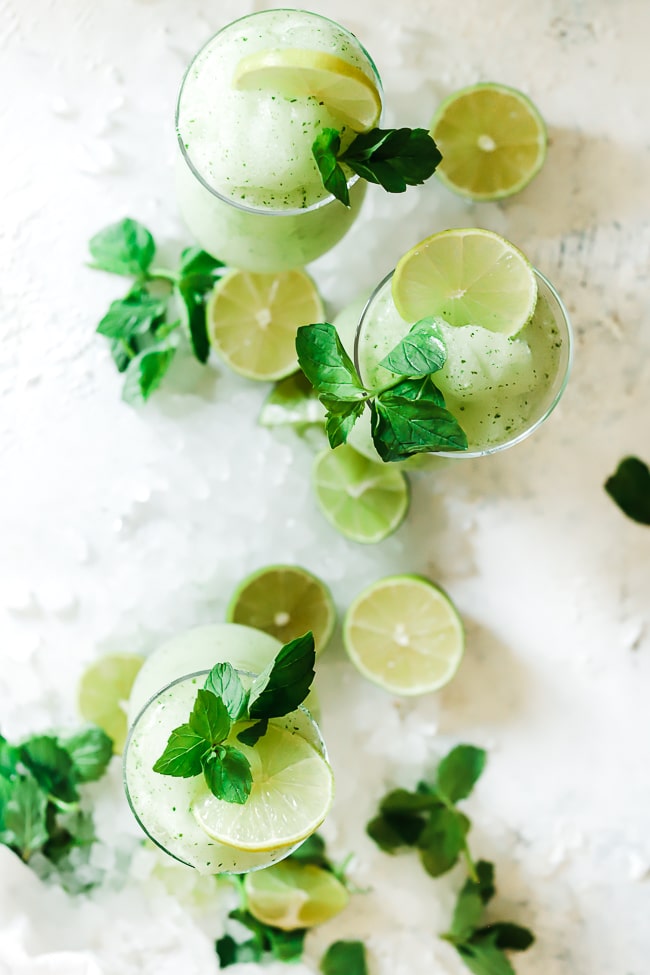 A flat lay of limeade slush. The glasses are surrounded by crushed ice, lime halves, and fresh mint.