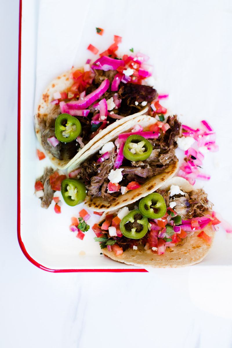 Three tacos topped with red pickled onions on a white tray