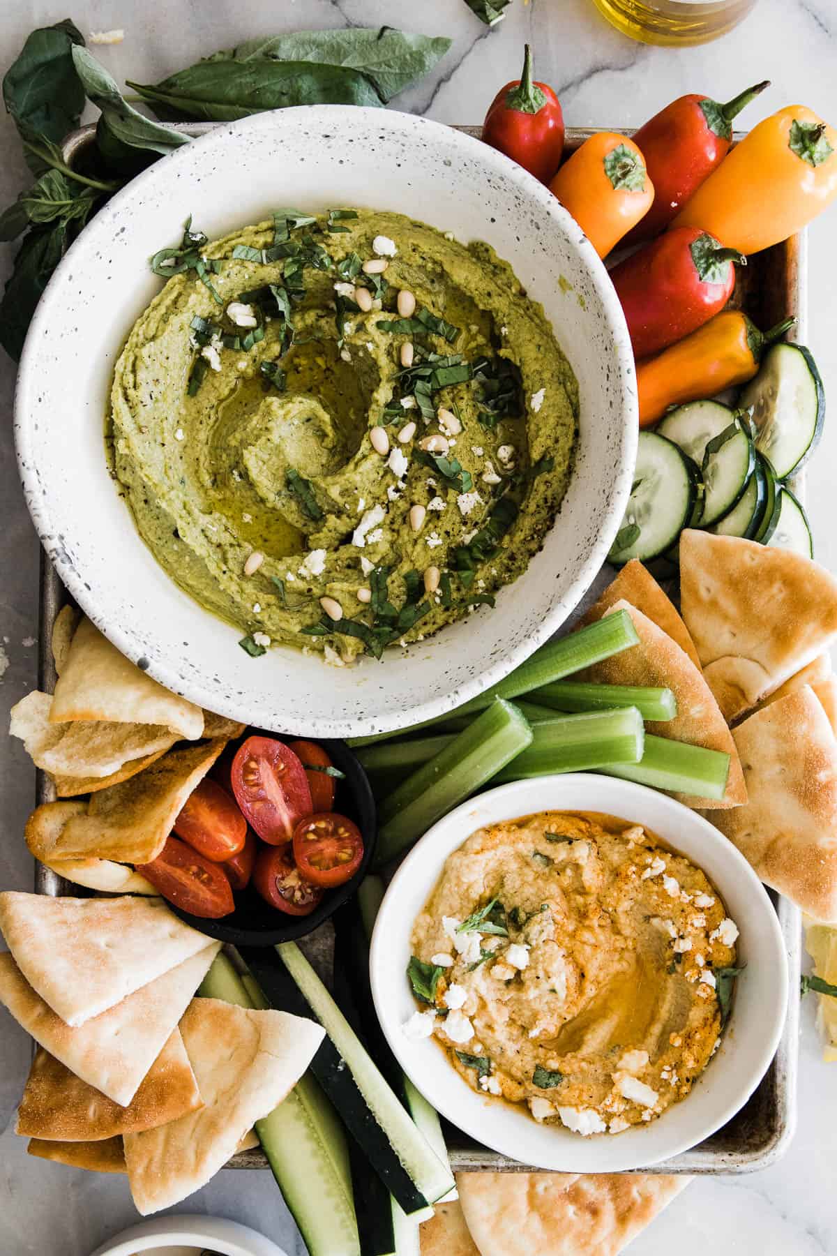 Bowl of green creamy pesto hummus with basil, vegetables, peppers and pita bread around it. 