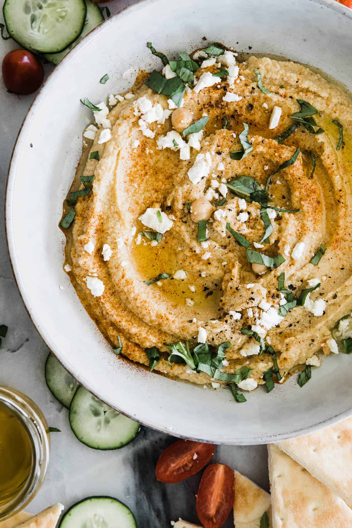 A bowl of healthy hummus drizzled with olive oil and sliced cucumbers and tomatoes on the side.