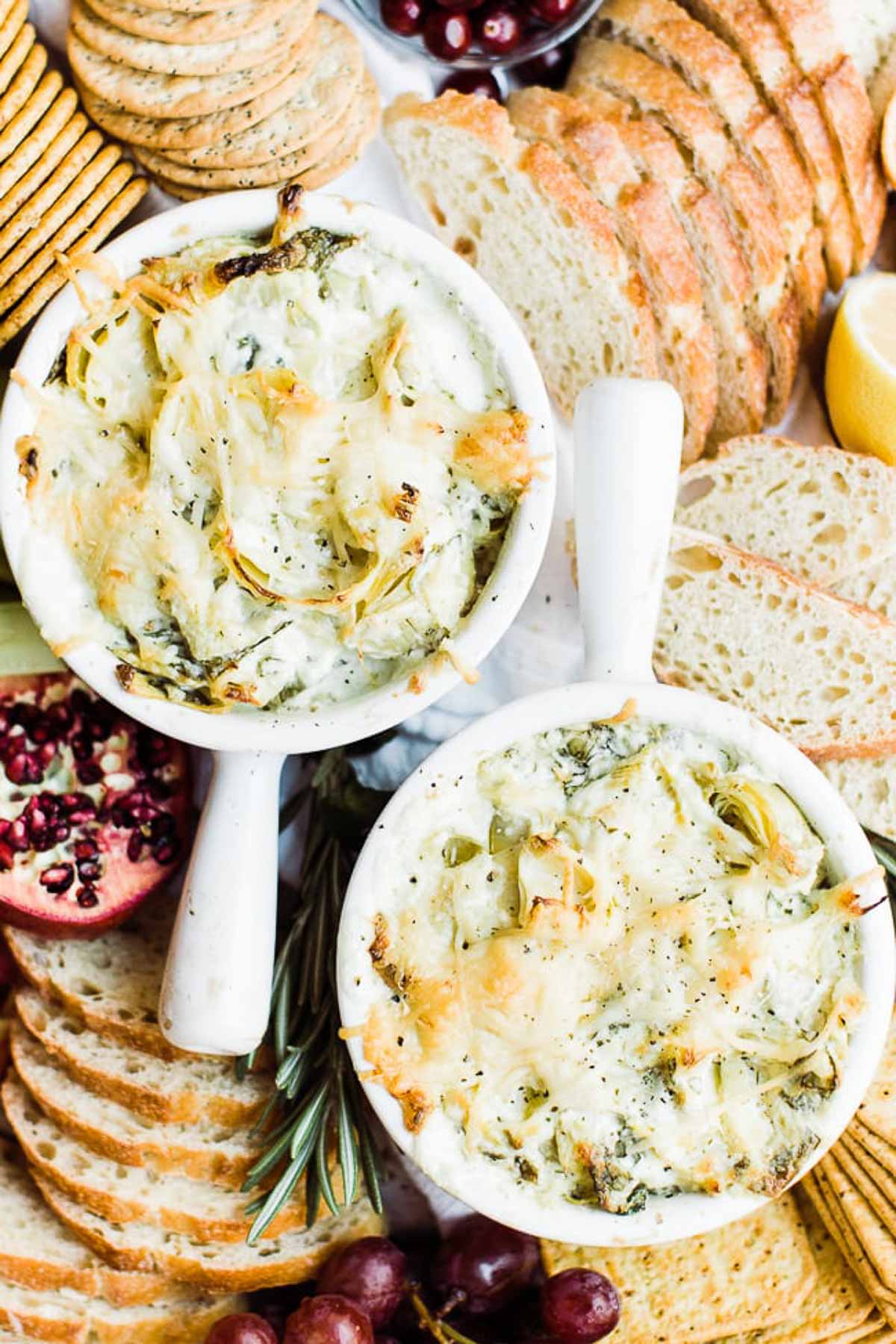 Two bowls of cheesy spinach dip on a tray surrounded by bread and crackers.