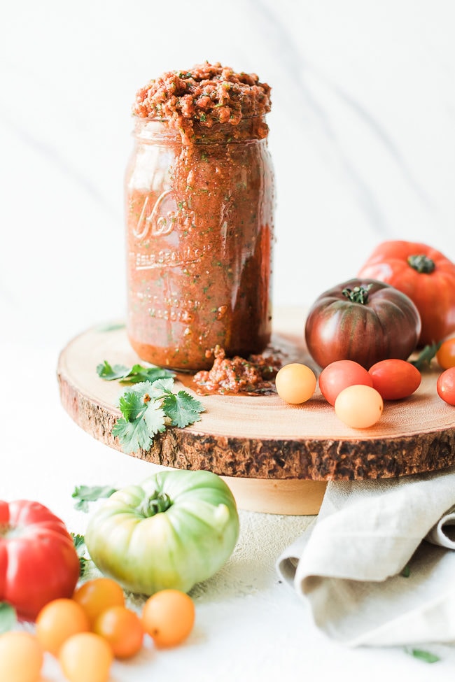 Chipotle salsa recipe in a mason jar, atop a wooden cake stand, surrounded by tomatoes.