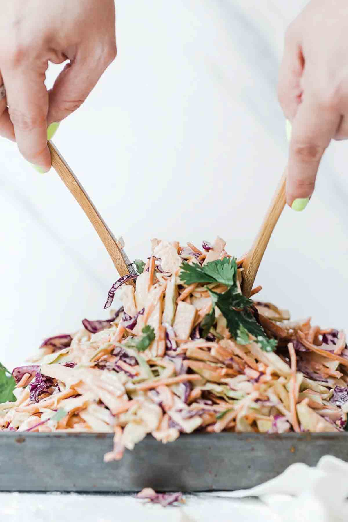 Apple cabbage slaw on the table with hands using spoons to lift out a serving.