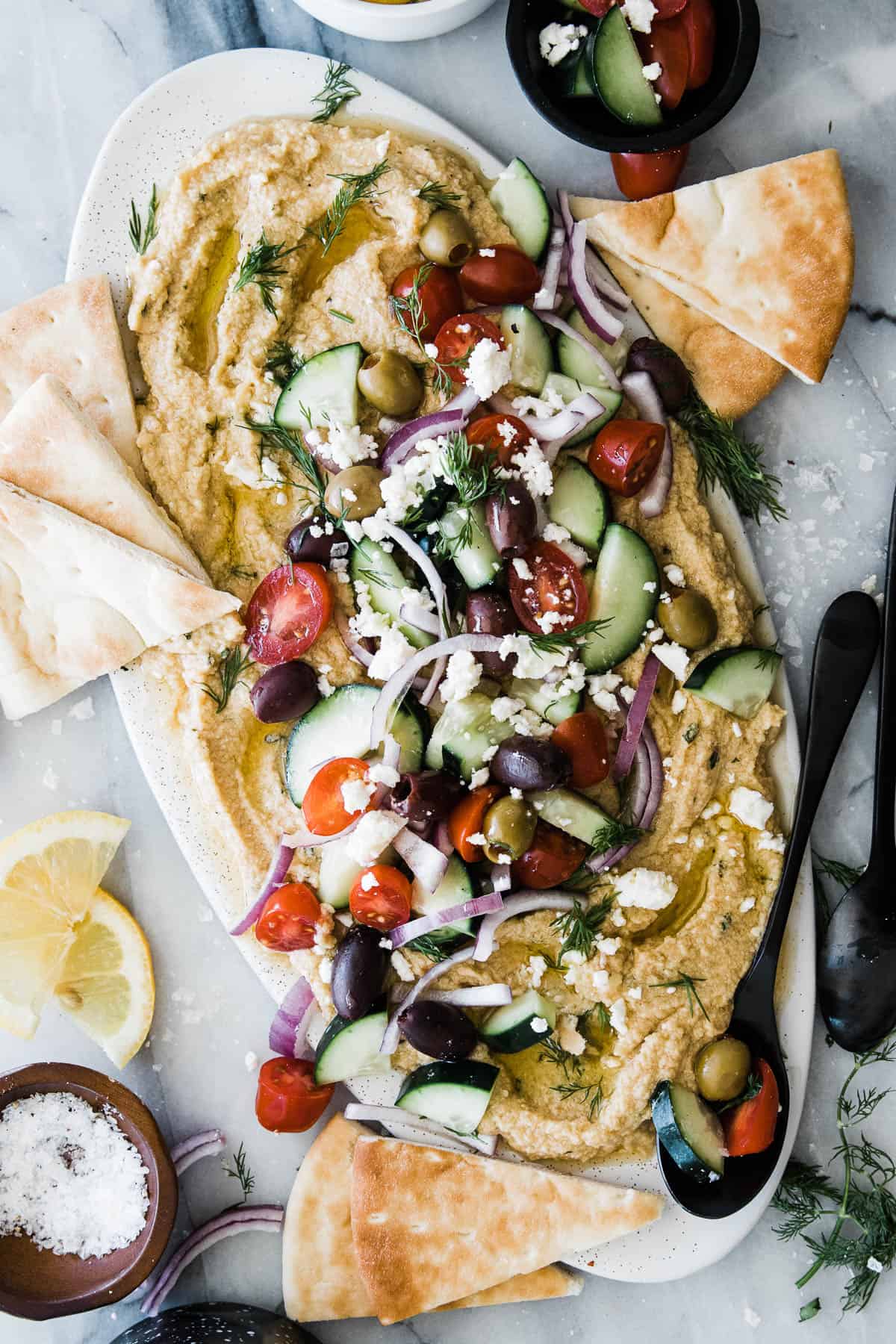 Platter of greek hummus loaded with vegetables and fresh herbs with pita bread on the side. 
