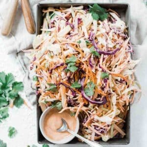 Homemade coleslaw in a metal tray. There is a cup of dressing to the side.
