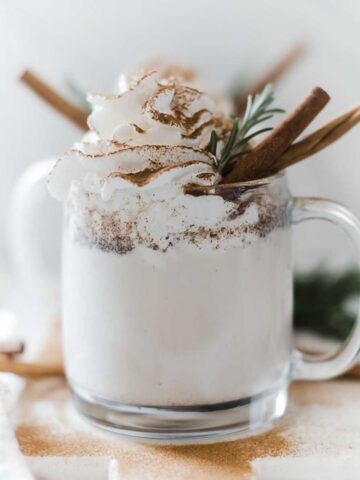 Pressure cooker warm milk and cinnamon in glass mugs, topped with whipped cream and garnished with rosemary and a cinnamon stick in a glass mug.