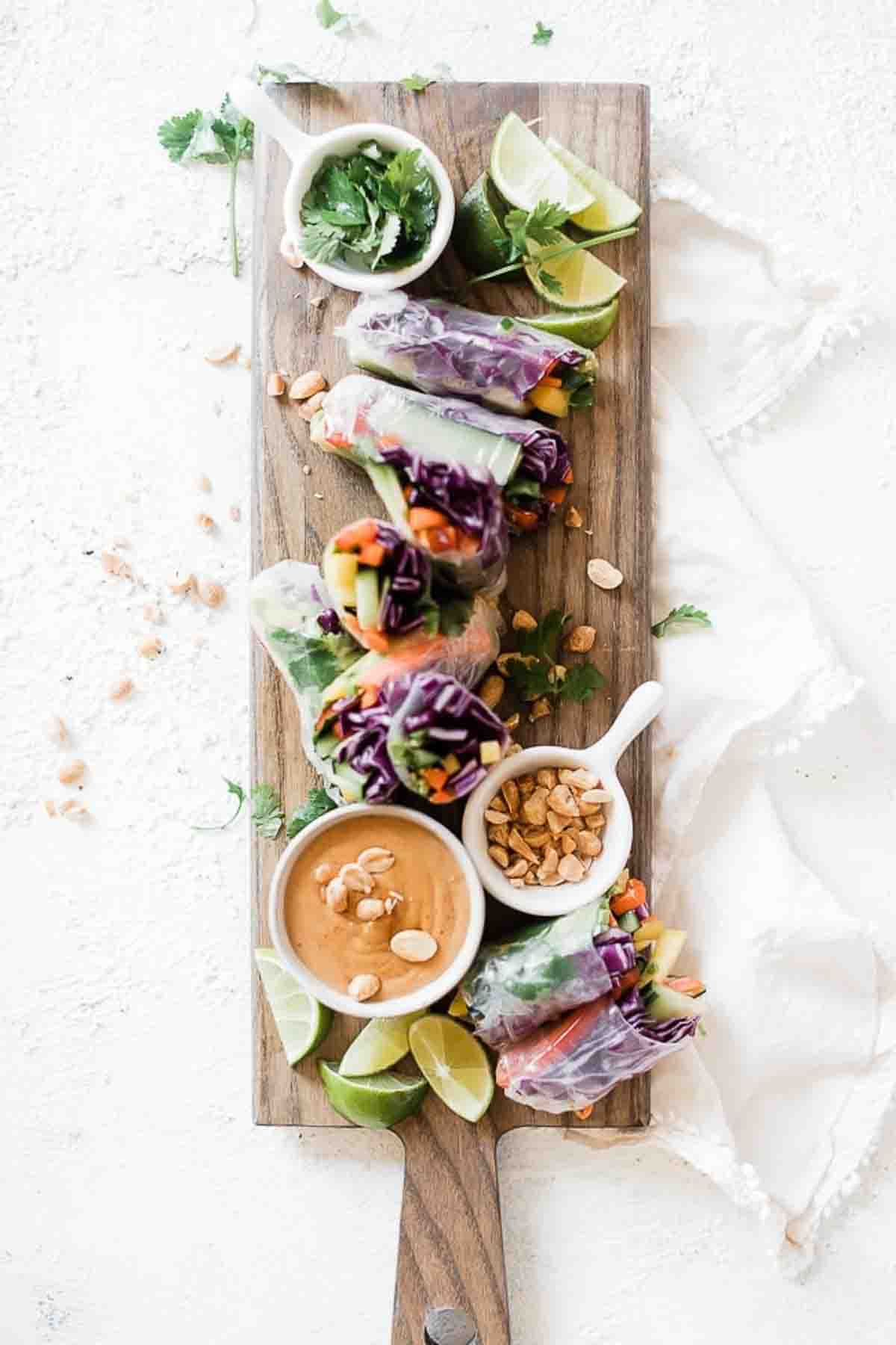 Rice paper rolls arranged on a wooden cutting board with bowl of peanut dipping sauce and peanuts to the side.