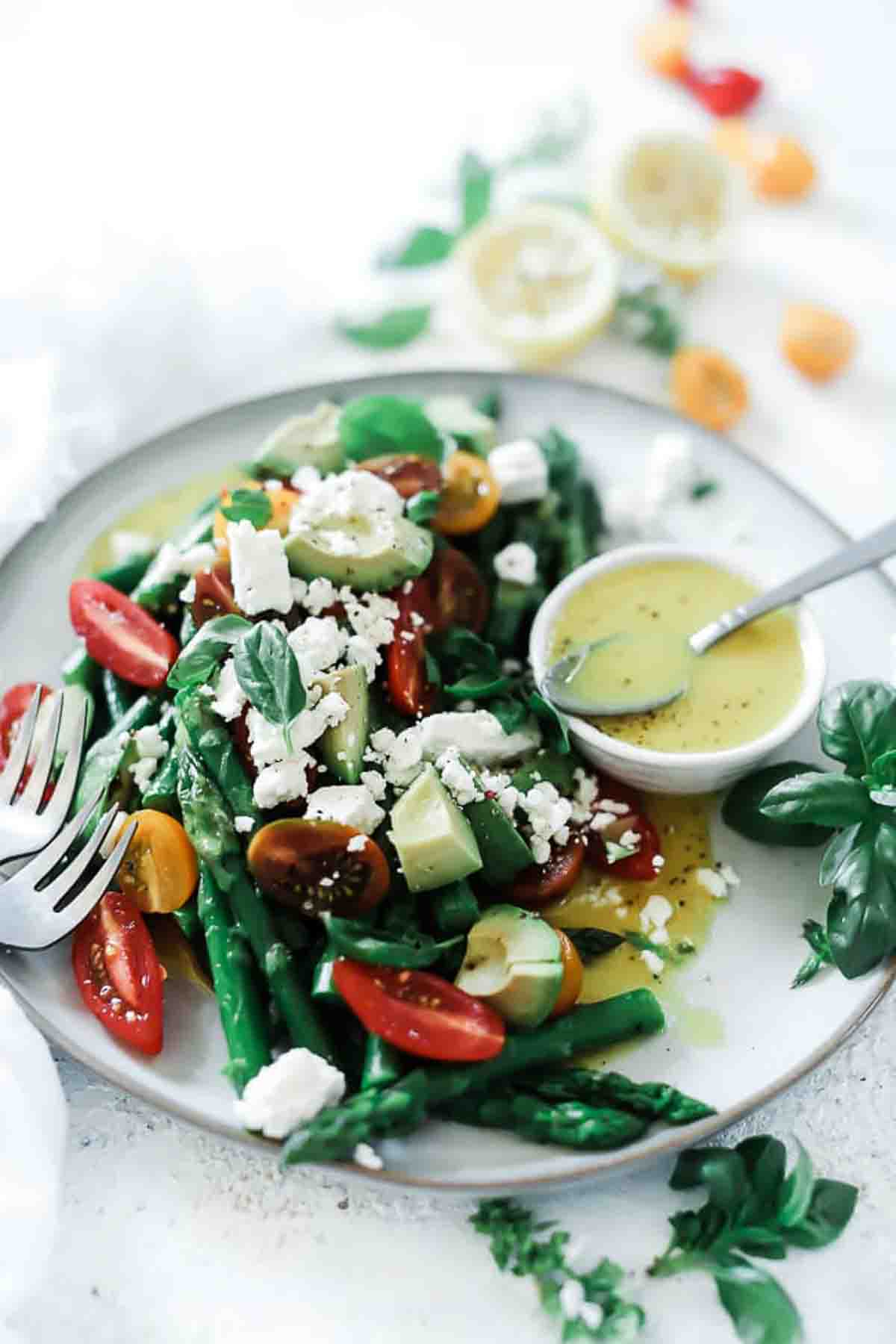A ¾ angle shot of asparagus salad on a white plate. The salad includes asparagus, tomato, avocado and feta cheese. It is garnished with basil and has a small bowl of dressing to the side.