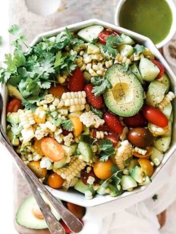 Summer corn salad in a grey bowl. IT is garnished with cilantro. There is a small bowl of dressing to the side and salad serves on the side of the bowl. The bowl is on top of a wooden cutting board with a white napkin to the side.