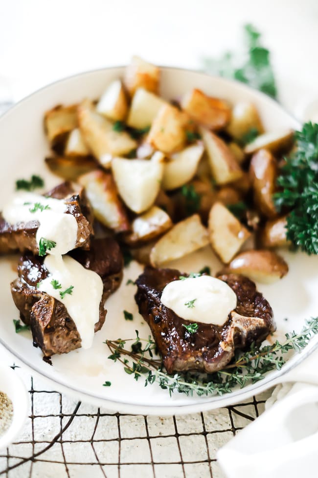 Lamb chops in a white braiser, topped with feta cream sauce. The braiser is filled with potatoes.