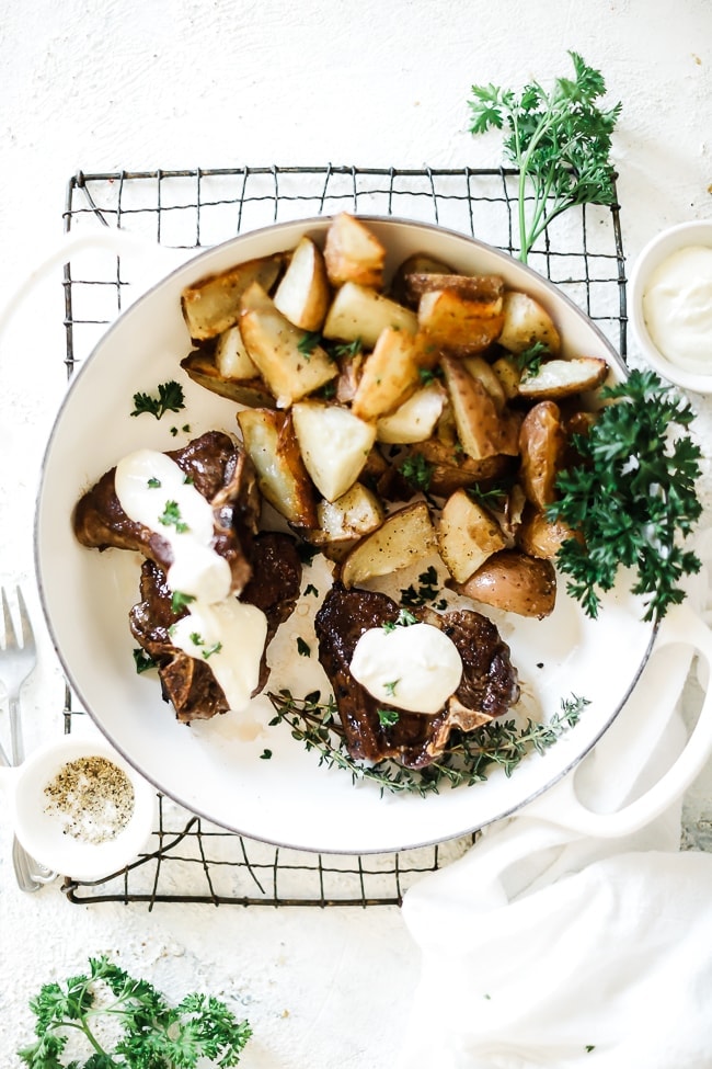 Lamb chops in a white brasier with roasted potatoes, topped with feta cream sauce. The dish has been garnished with parsley.