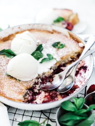 Berry Cobbler recipe in a white braiser. The pan is set atop a wire cooling rack and is garnished with mint and ice cream.