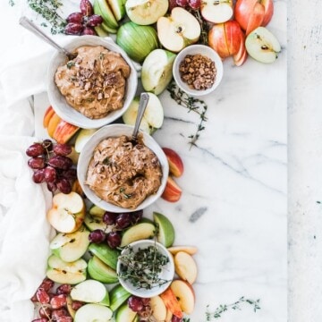 A marble platter filled with green and red apples, grapes, and two bowls of toffee apple dip.