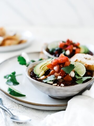 A 3/4 view of black bean soup garnished with avocado, pico, feta cheese and lime.