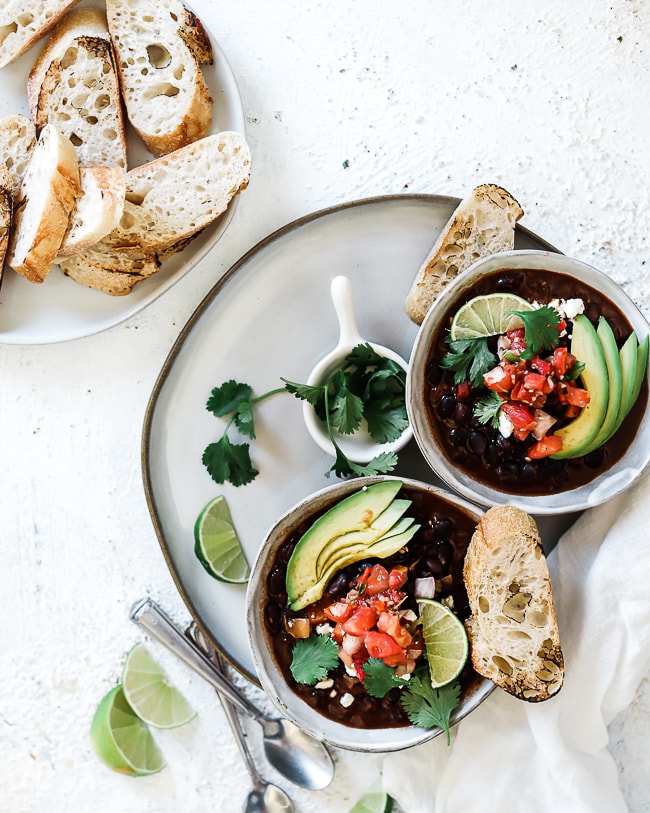 Two bowls of Panera black bean soup atop a grey plate. There is a pinch bowl of cilantro, a baguette slice, and a lime on the plate. There are spoons and a white napkin to the side. There is also a platter of sliced baguettes.