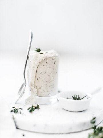 Homemade ranch dip in a glass jar with a spoon leaning against it. There is a small bowl of herbs to the side.