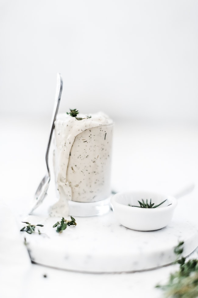 Homemade ranch dip in a glass jar with a spoon leaning against it. There is a small bowl of herbs to the side.