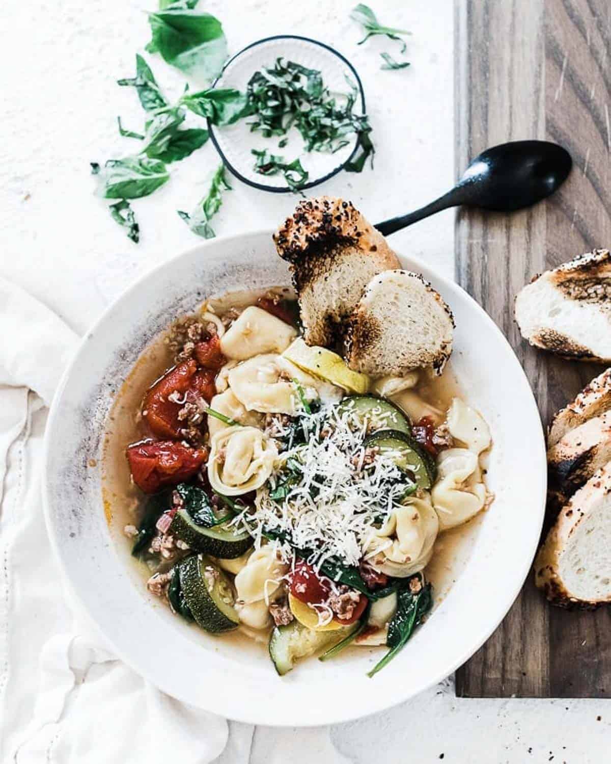 Sausage Tortellini soup in a bowl on the table with a spoon to the side and bread on a cutting board.