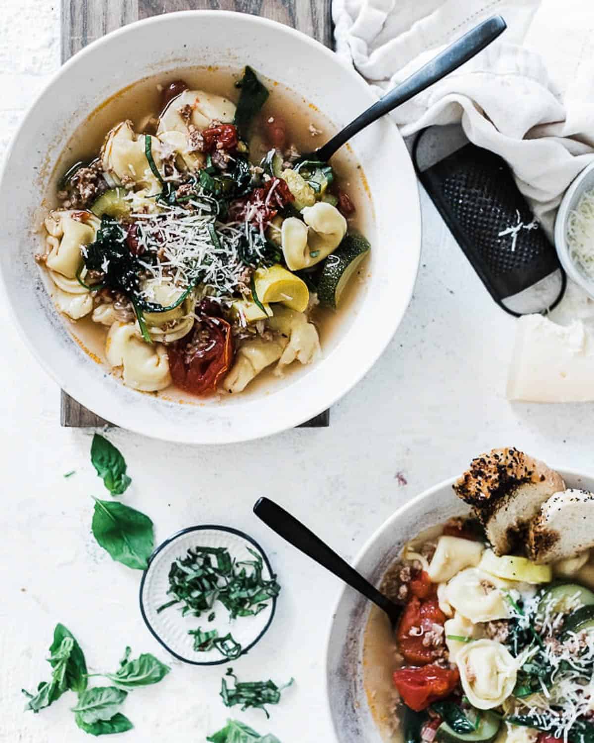 Italian sausage tortellini soup in white bowls with black spoons sticking out and there is a small plate of basil to the side.