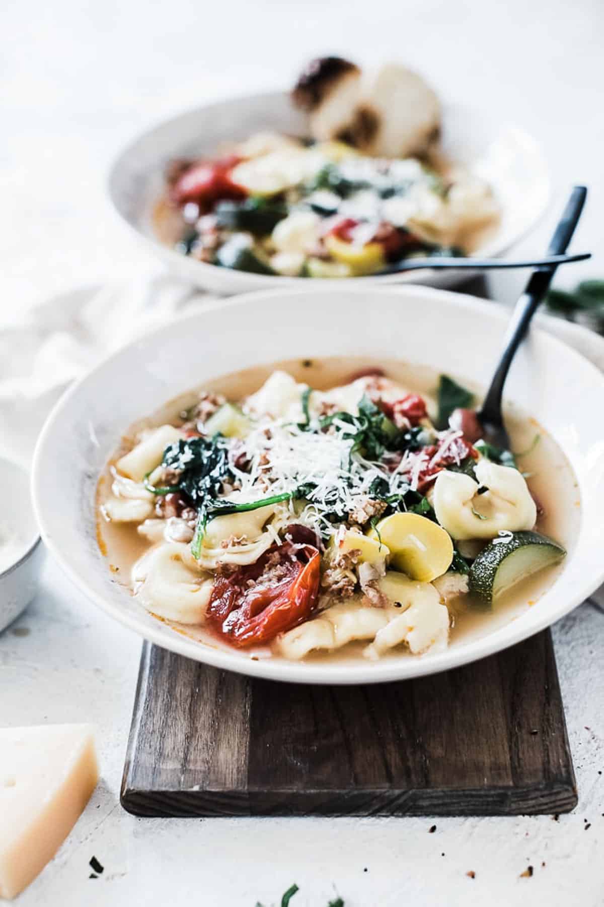 Italian sausage tortellini soup in white bowls atop a wooden cutting board. 