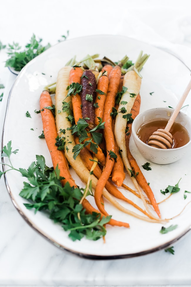 A ¾ angle shot of pressure cooker carrots in a white platter. There is a bunch of parsley to the side and a small bowl of honey with a honey dipper in it.